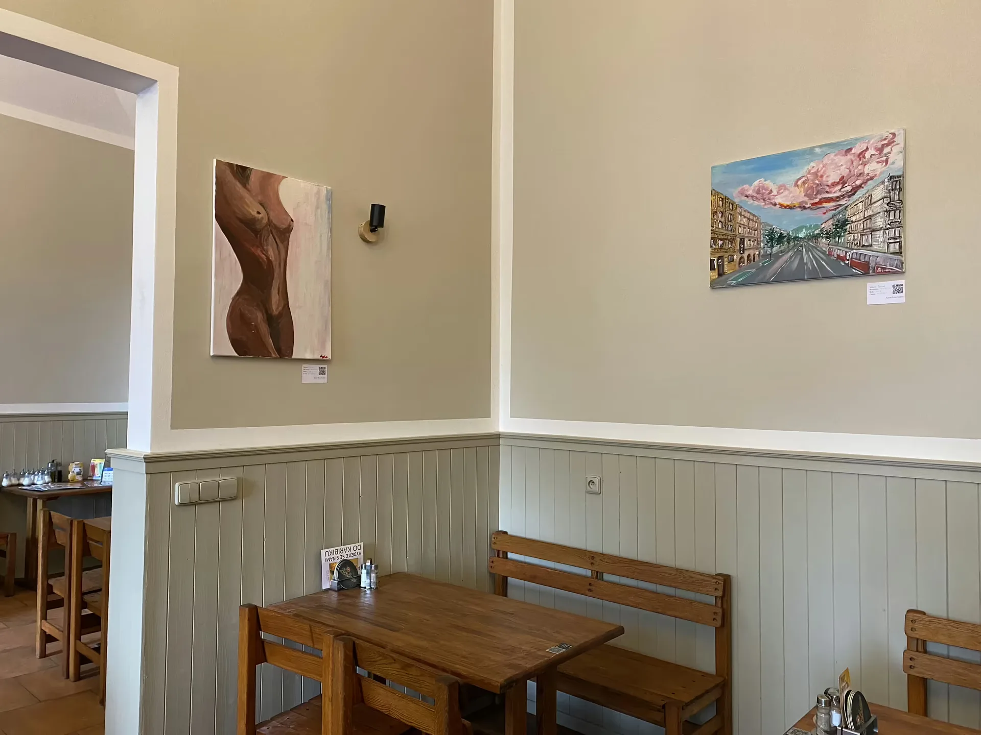 Two paintings by Ivan Anikin installed on the cream walls of a Prague cafe above wooden tables.