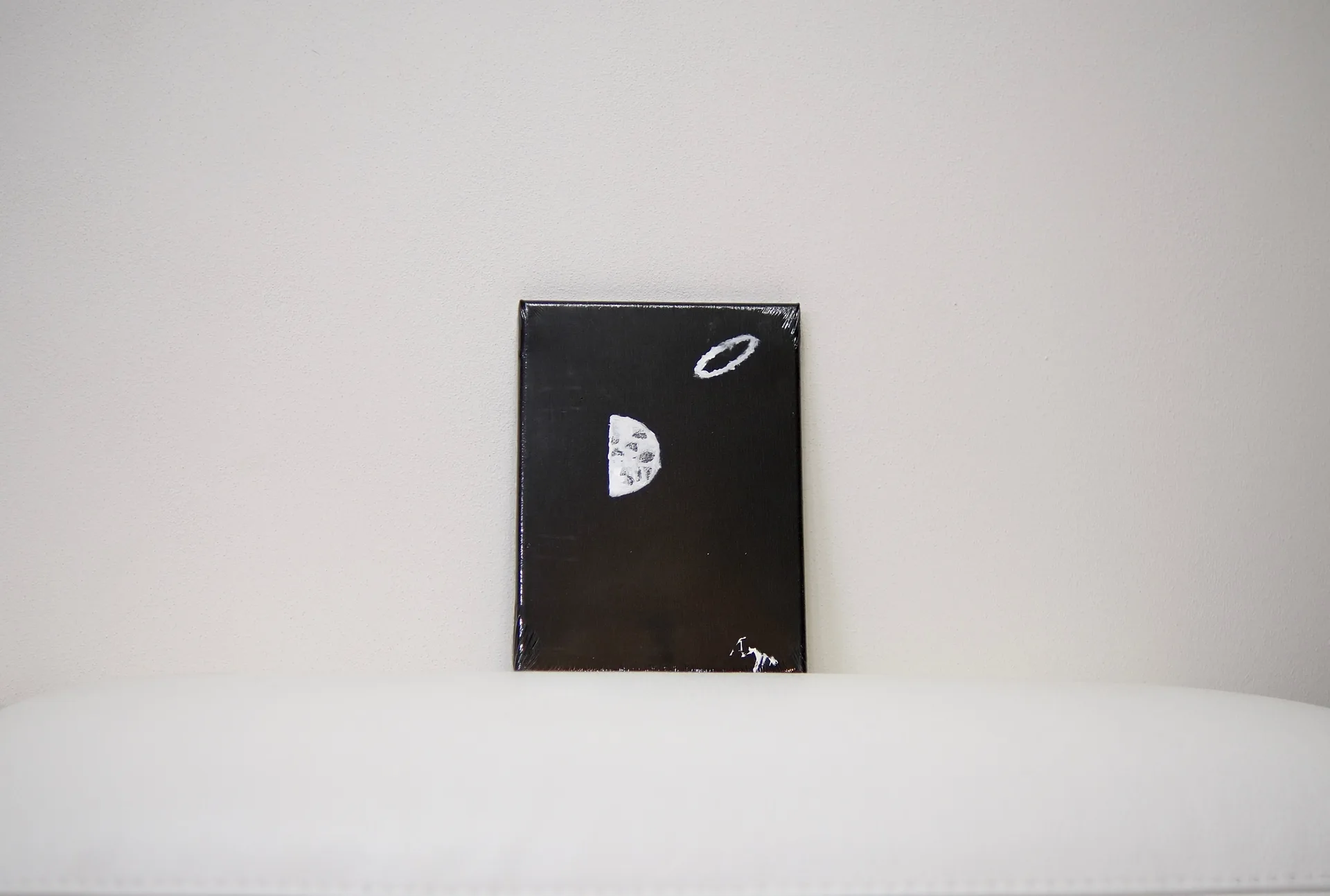 The small black canvas standing on a white cushioned surface against a pale wall, dwarfed by the empty space around it.